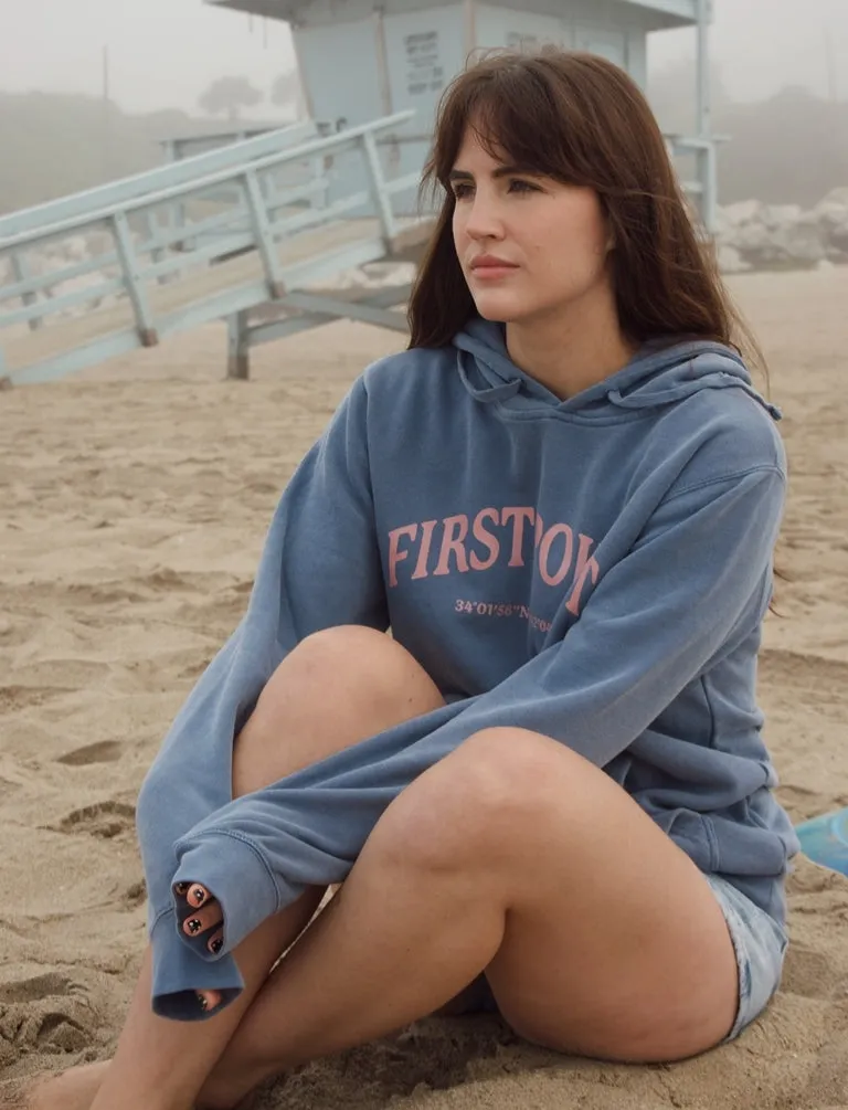 A surfer in a First Point hoodie sitting on foggy sand below a lifeguard tower