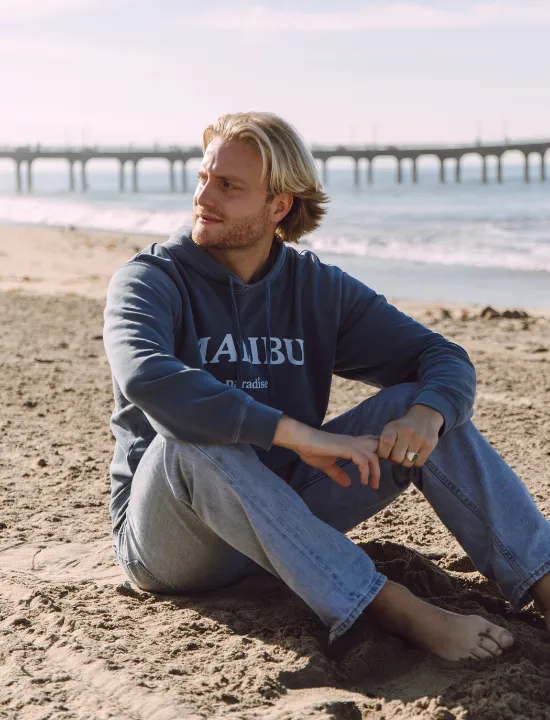 A man in a Malibu hoodie sitting on the beach by the pier