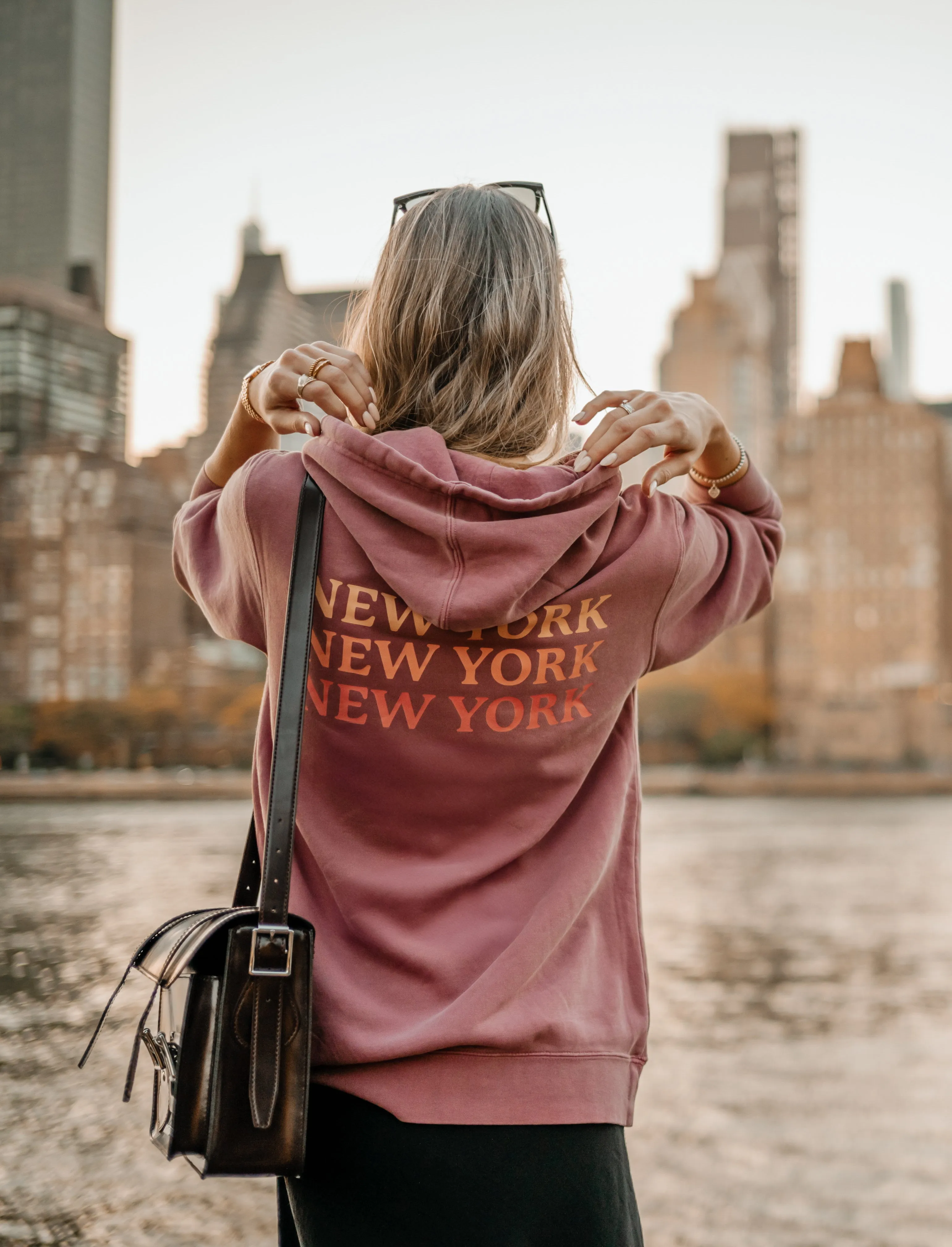 Back of a mauve New York New York New York hoodie, worn on the East River with the Manhattan skyline behind