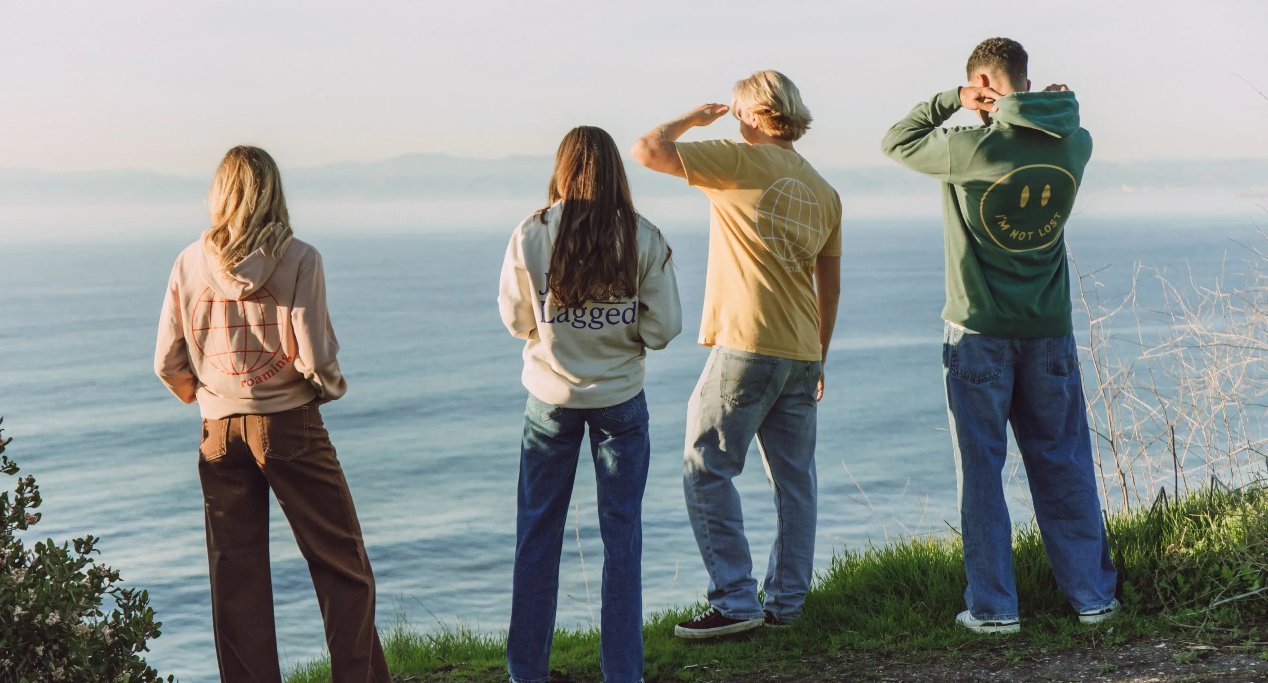 Four friends in Paradise & Main hoodies and tees watching the ocean from a bluff at golden hour